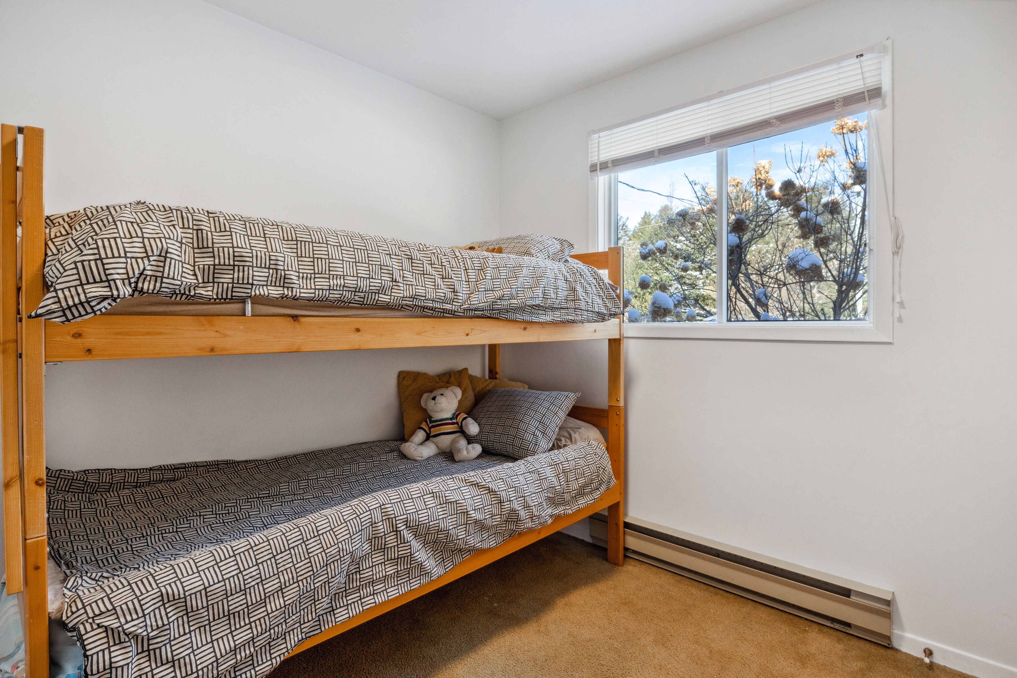 A bunk bed with patterned bedding, a teddy bear, and a window with a view of trees.