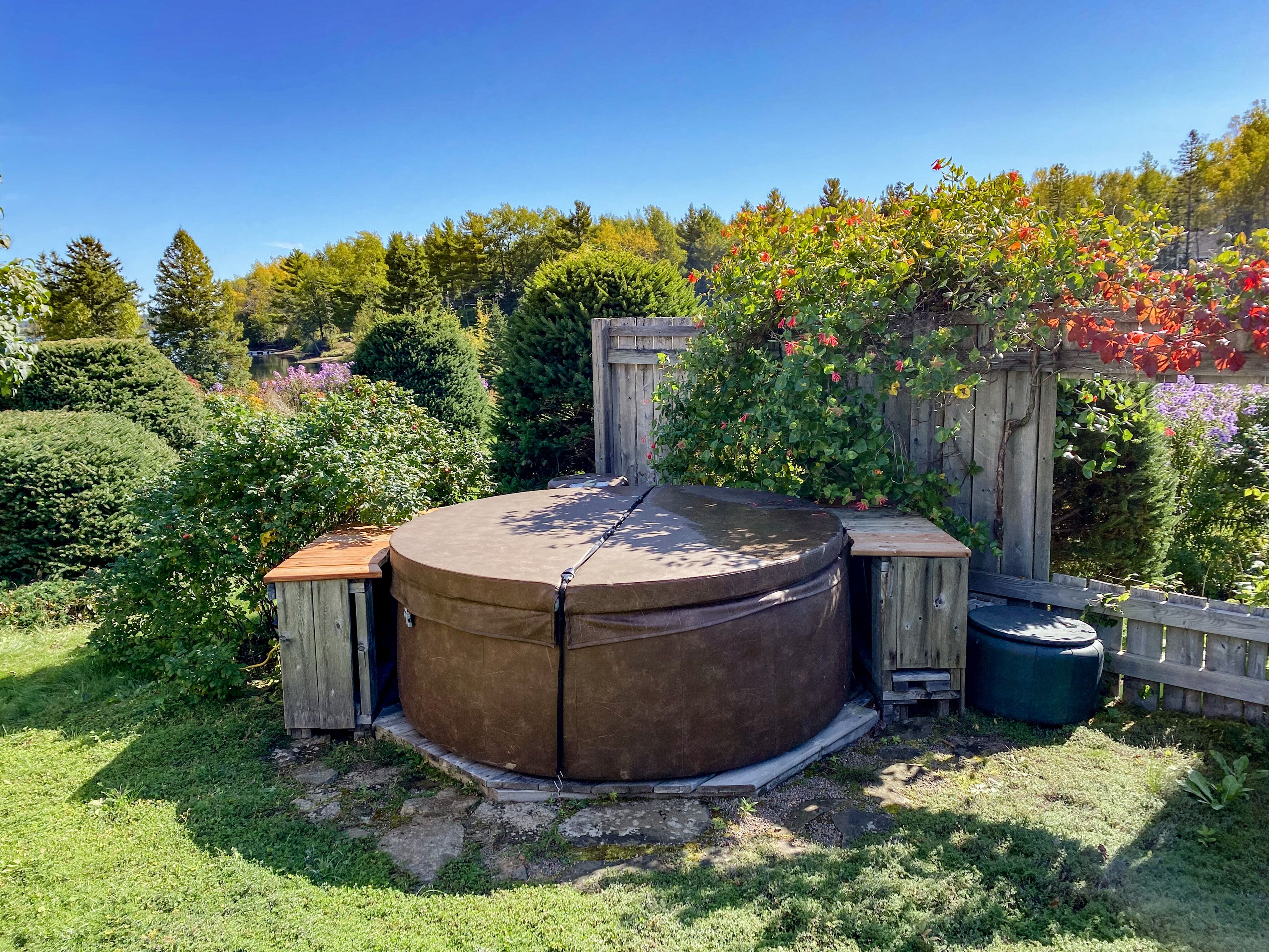 A circular outdoor hot tub with wooden benches on either side, surrounded by lush greenery and a wooden fence.