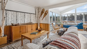 A modern living room with a wooden table, a sofa, and a large window offering a view of trees.