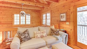 A wooden cabin interior with a sofa, pillows, a ceiling fan, and large windows. - 'Toccoa Waterfront Hideaway:' Hot Tub & Fire Pit! (Mineral Bluff)