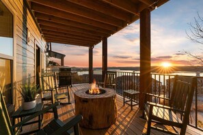 A wooden deck with a fire pit, outdoor seating, and a view of the sunset over a body of water.