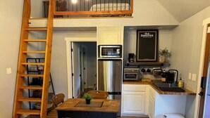 Een compacte keuken met een koelkast, oven en magnetron. Er staat een houten tafel met stoelen en een kleine potplant. Een houten ladder staat tegen de muur en een prullenbak bij de deur.