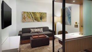 A modern hotel room with a flat-screen TV, a gray sofa with a patterned pillow, a wooden side table, a desk with a lamp, and two artworks on the walls. - Springhill Suites By Marriott Ellijay (Ellijay)
