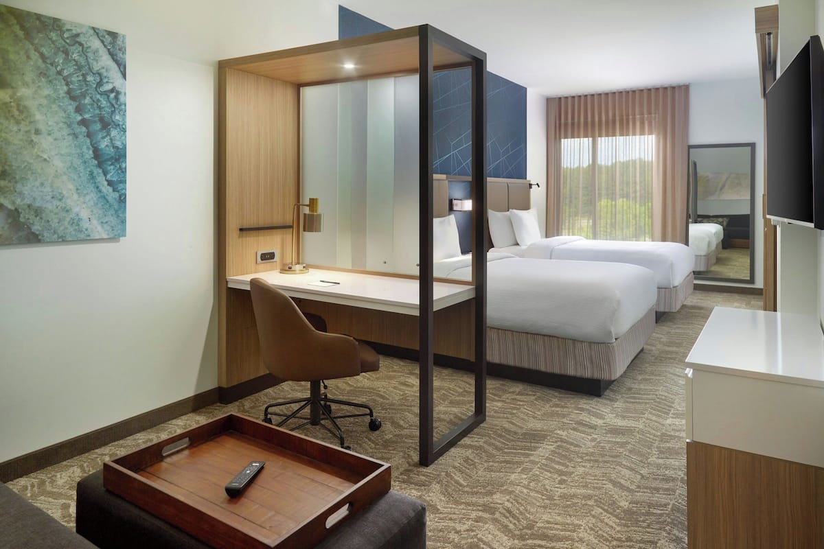 A hotel room with a desk, two beds, a chair, a TV, and a large window.