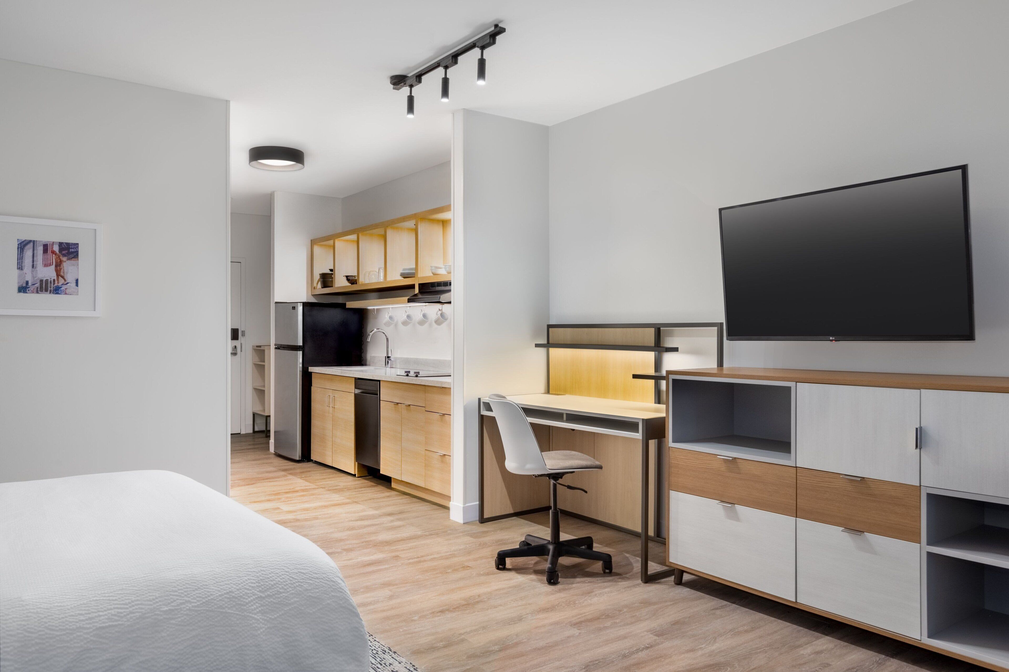 A modern hotel room with a bed, a desk, a TV, and a kitchenette.