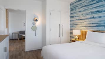 A hotel room with a large bed, a nightstand with a lamp, a wall with a blue abstract painting, a door leading to another room, and a small seating area with a chair and table.