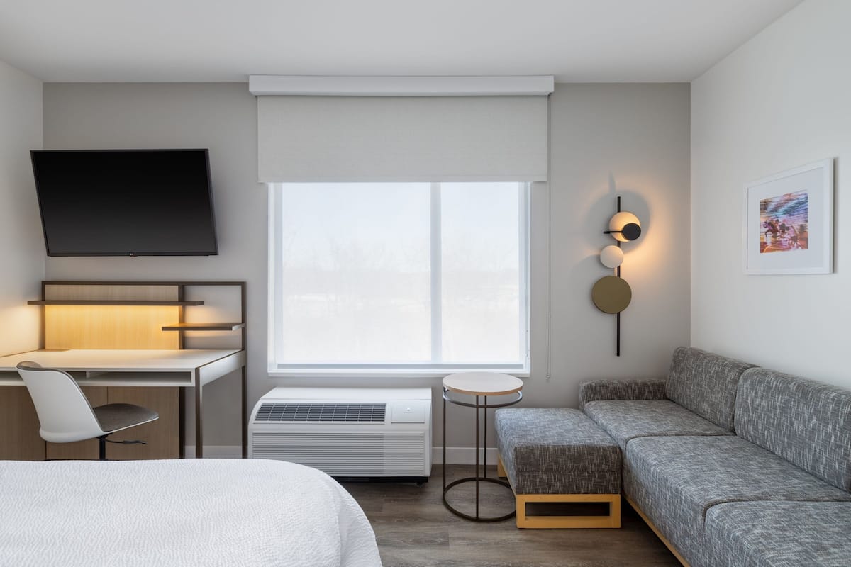 A modern hotel room with a bed, a desk with a chair, a sofa, a TV, a window with blinds, and a small table.