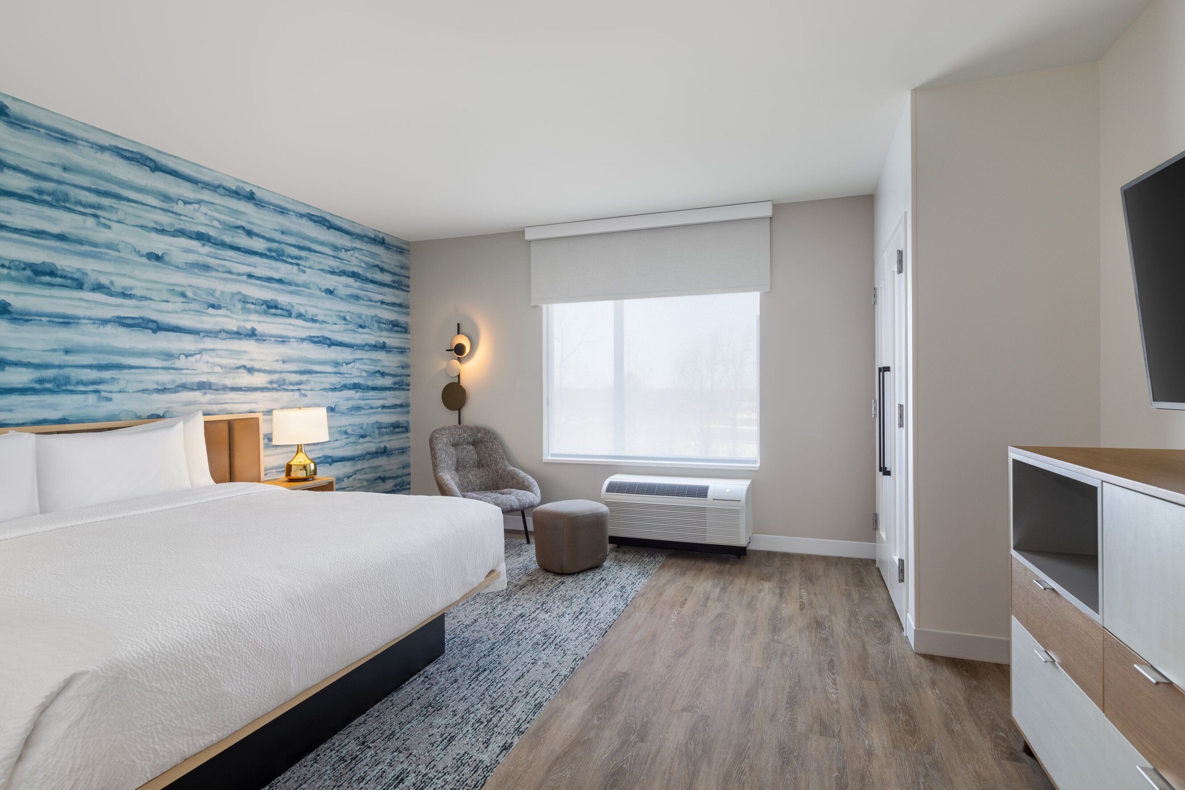 Una habitación de hotel moderna con una cama grande, un área de descanso con una silla, un televisor y una ventana con persianas.