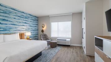 Una habitación de hotel moderna con una cama grande, un área de descanso con una silla, un televisor y una ventana con persianas.