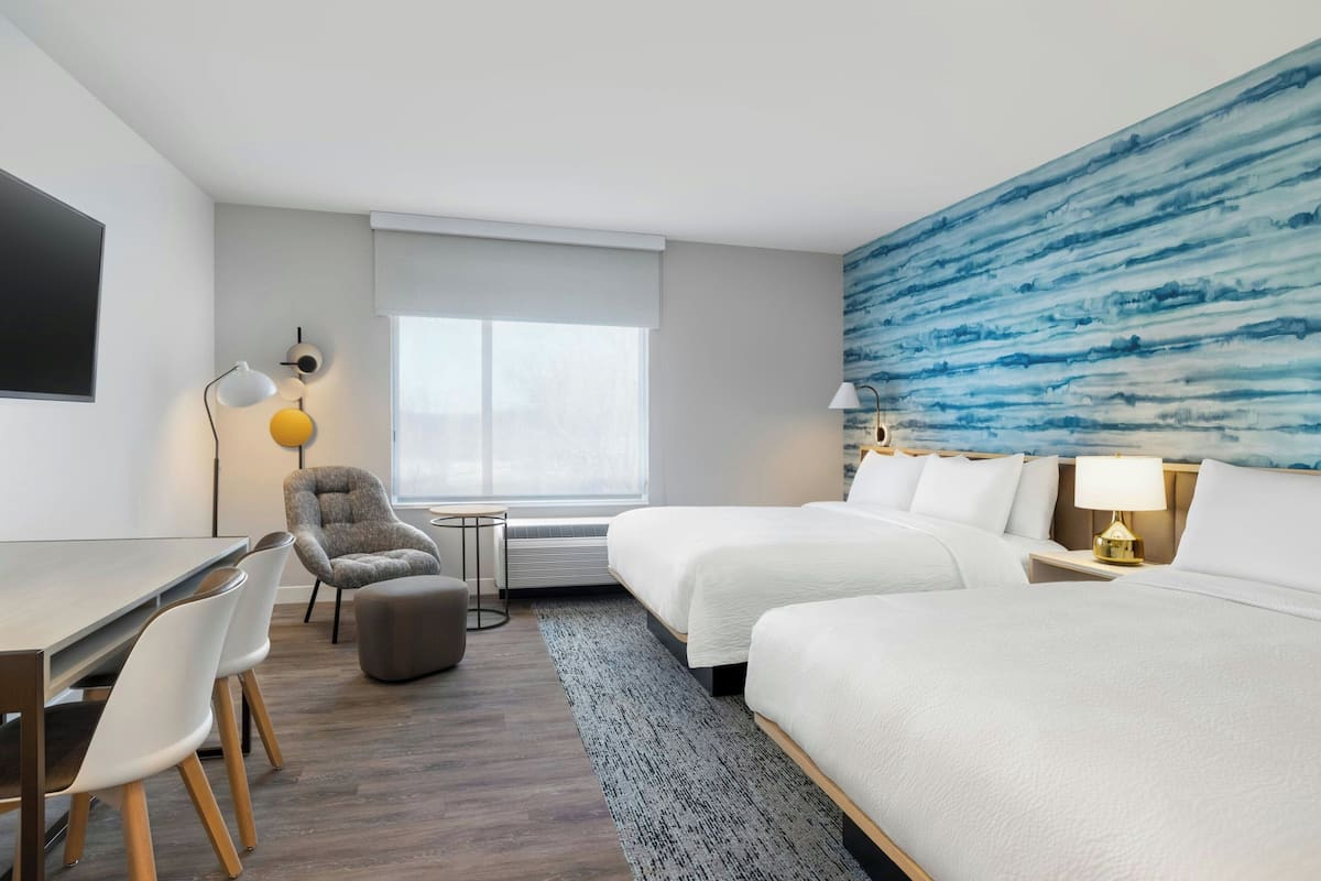 A hotel room with a bed, a desk, a chair, a lamp, and a large abstract painting on the wall.