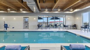 Ein Schwimmbecken mit Liegestühlen und Sonnenschirmen, Holzbalken an der Decke und Blick auf ein Gebäude im Hintergrund.