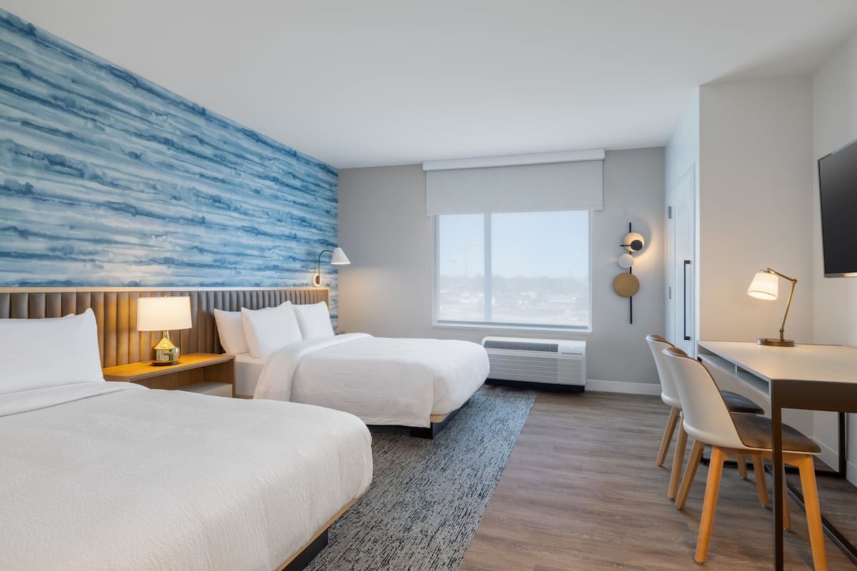A hotel room with two beds, a desk, chairs, a TV, and a large window.