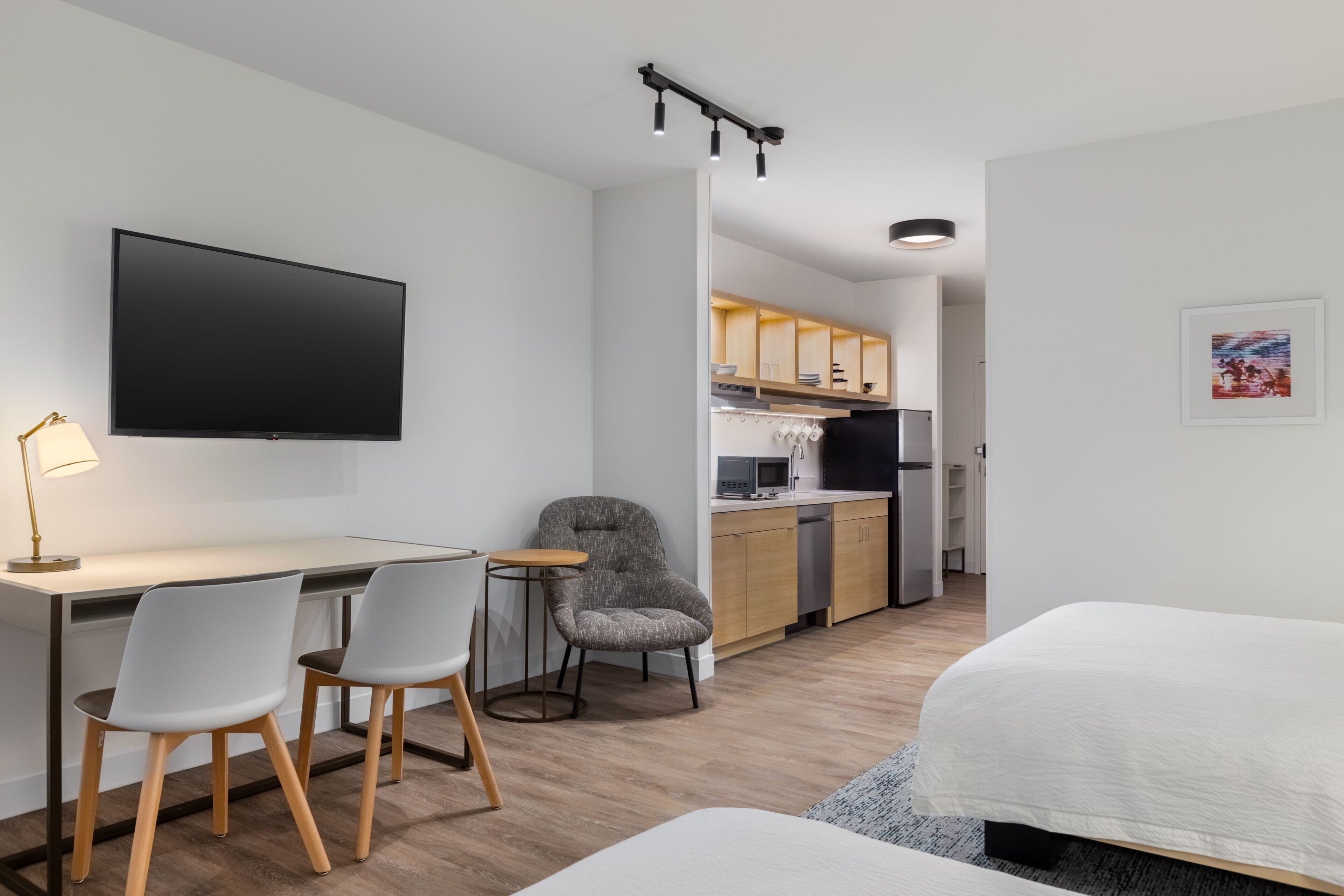 A modern hotel room with a desk, two chairs, a bed, a TV, and a kitchenette in the background.