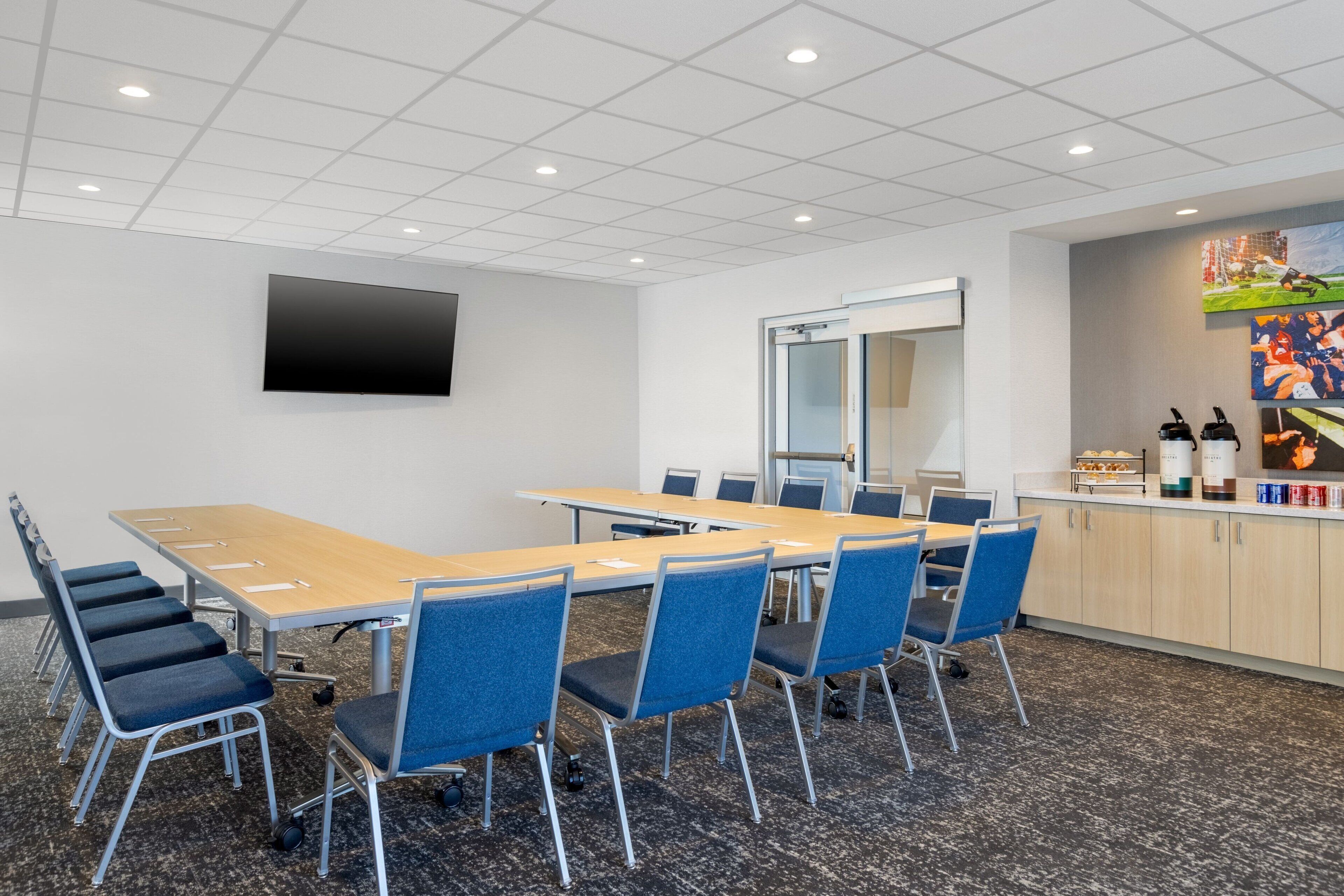 Une salle de conférence avec une longue table, des chaises bleues, un téléviseur à écran plat et une porte en verre donnant sur une autre pièce.