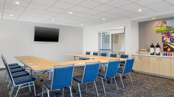 A conference room with a long table, blue chairs, a flat-screen TV, and a glass door leading to another room.