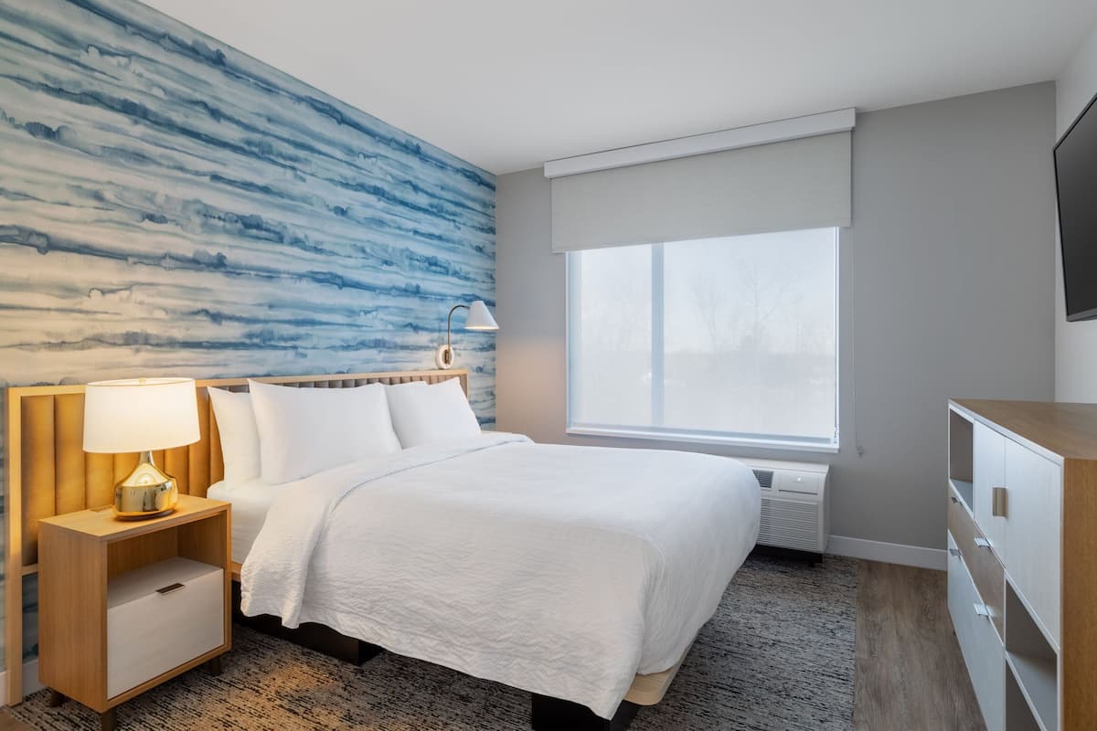 A modern hotel room with a large bed, a nightstand with a lamp, a dresser, and a large window with a view.
