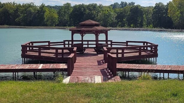 Ein hölzernes Pavillon auf einem Steg über einem See, umgeben von üppigem Grün.