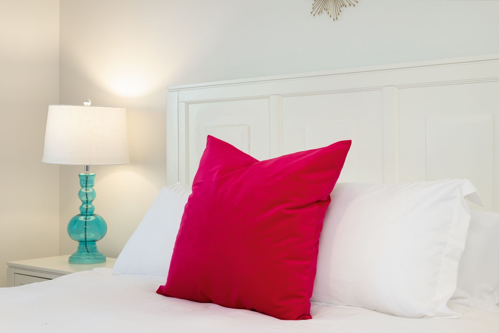 A neatly made bed with white and pink pillows, a teal lamp on a bedside table, and a white headboard.