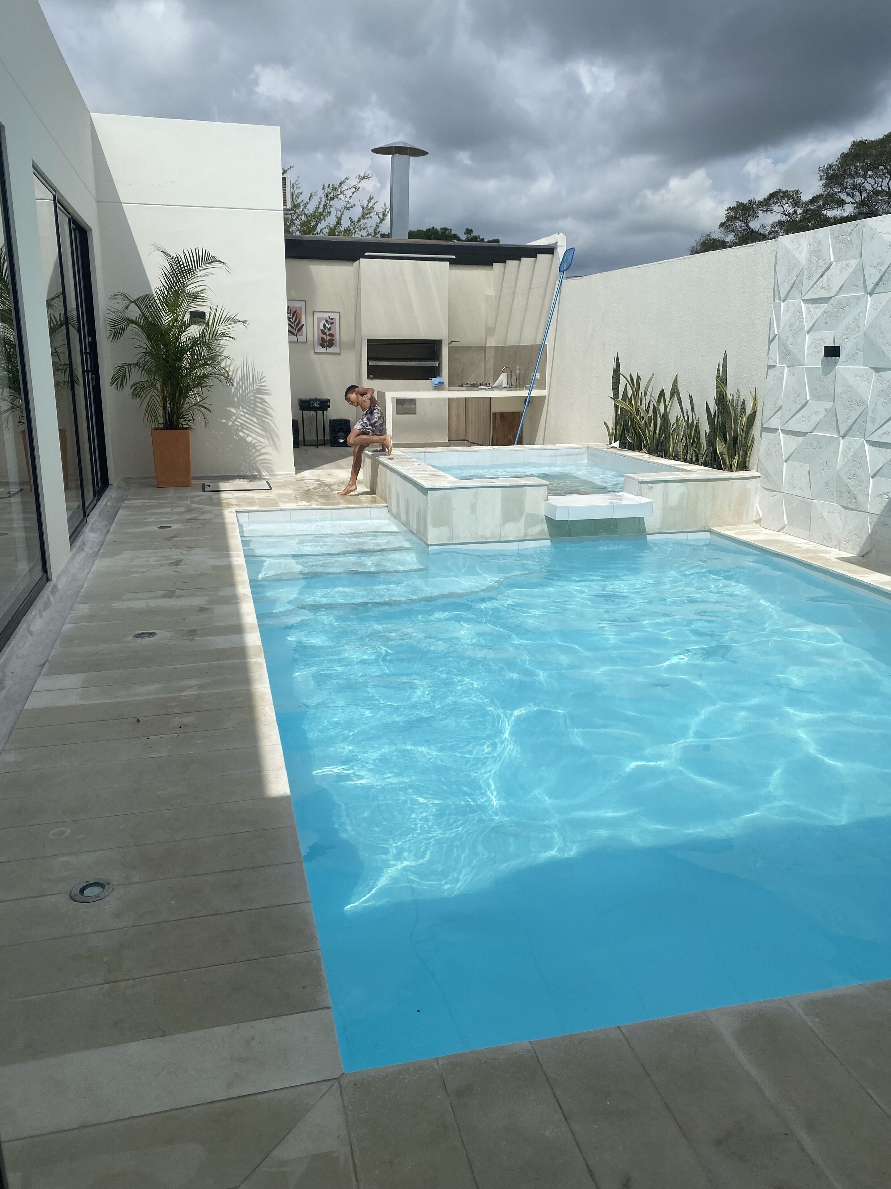 A modern outdoor swimming pool with a clear blue water surface, surrounded by a white wall with geometric patterns and a potted plant.