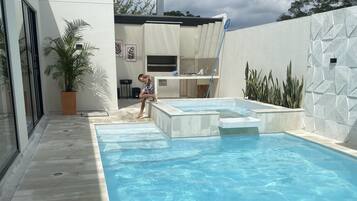 A modern outdoor swimming pool with a clear blue water surface, surrounded by a white wall with geometric patterns and a potted plant.