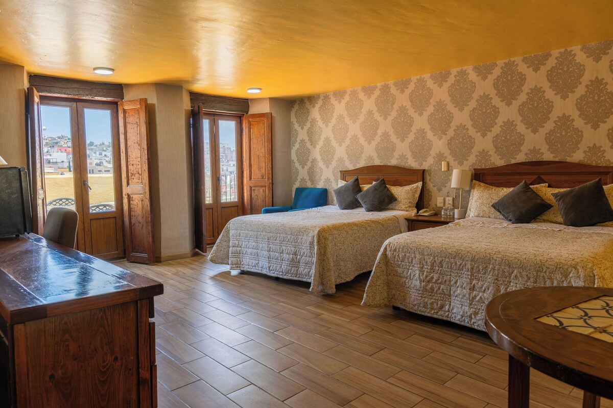 A hotel room with two beds, a wooden desk, and a view of the city.