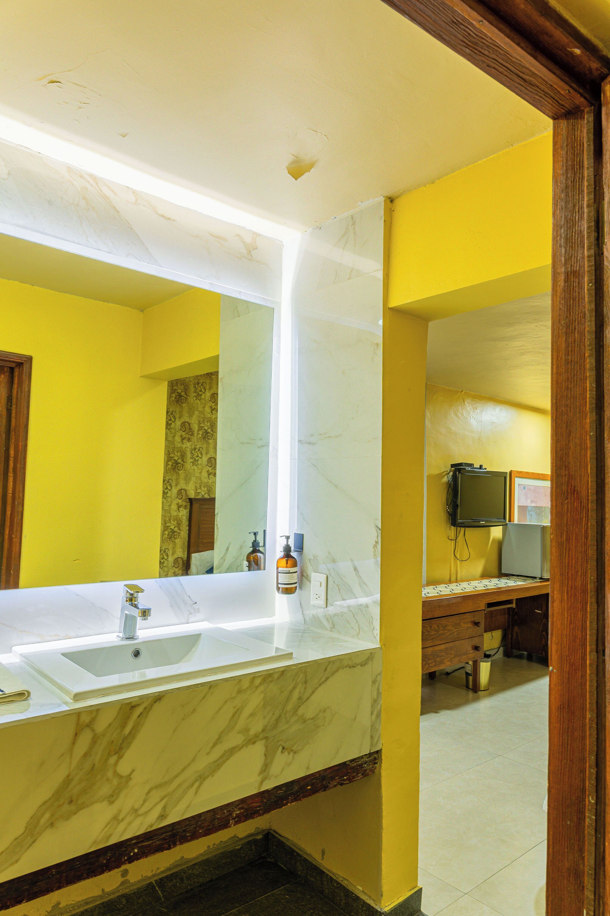 A bathroom with a marble sink, yellow walls, a mirror, a television, and a wooden cabinet.