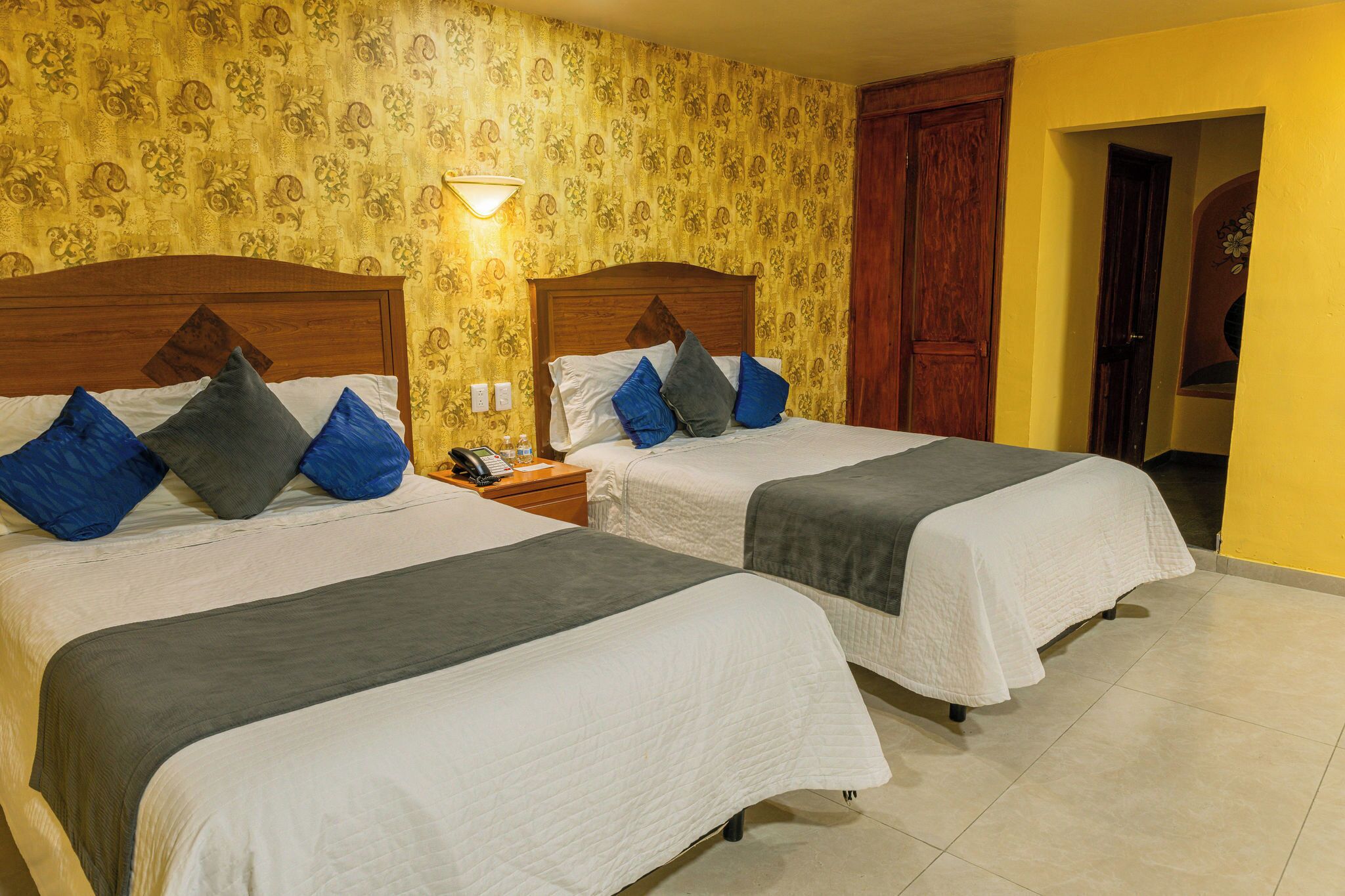 A hotel room with two beds, a nightstand, a lamp, and a patterned wallpaper.