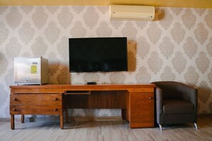 Une pièce comprenant un bureau en bois, un téléviseur fixé au mur, une chaise et un climatiseur.