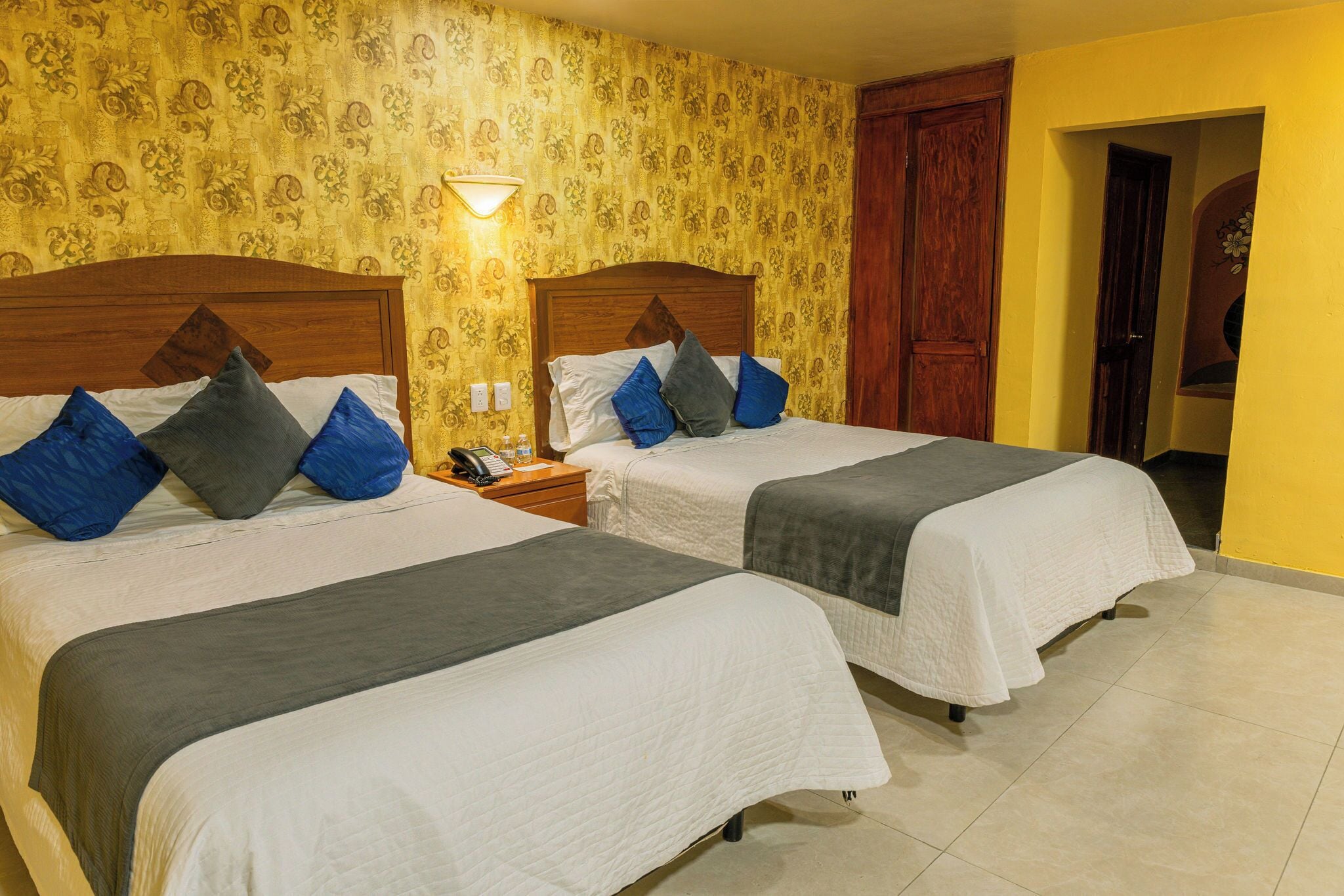 A hotel room with two beds, a nightstand, and a wall lamp.