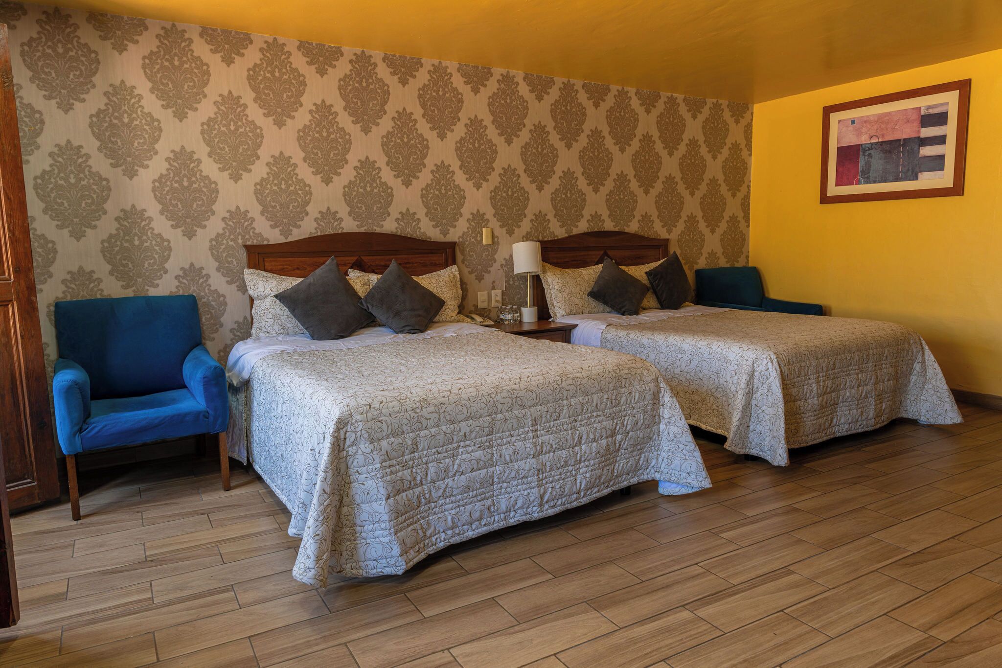 A room with two beds, a blue armchair, a bedside table with a lamp, and patterned wallpaper.