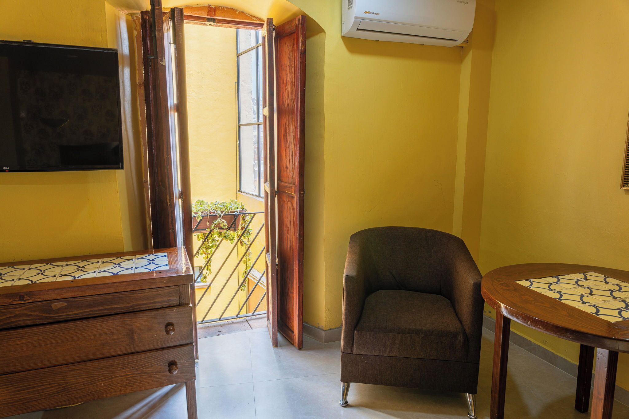 A room with a wooden dresser, a chair, a small table, a TV mounted on the wall, and an air conditioning unit.