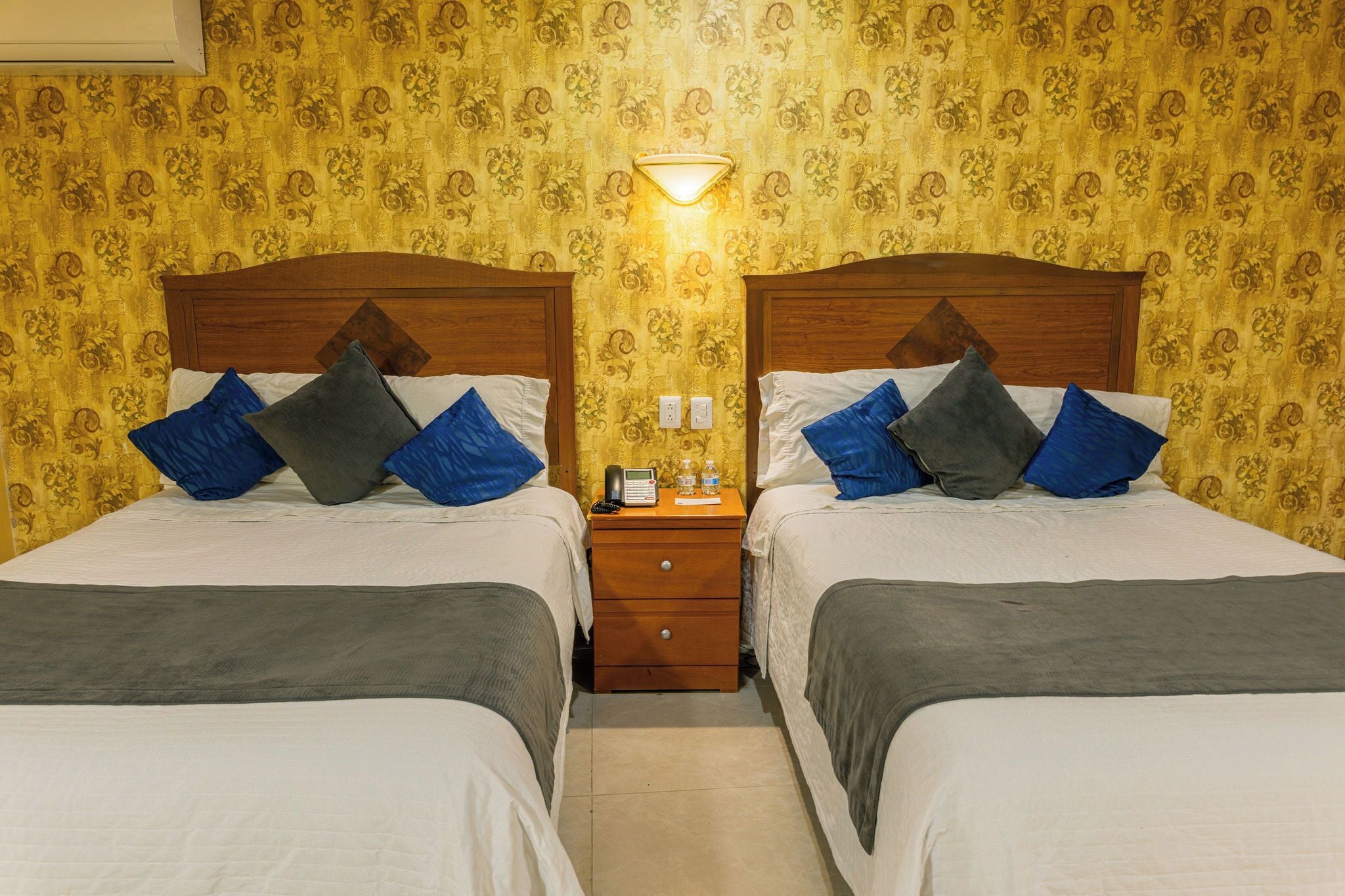 Habitación doble con dos camas, una mesita de noche, una lámpara y un papel tapiz estampado.