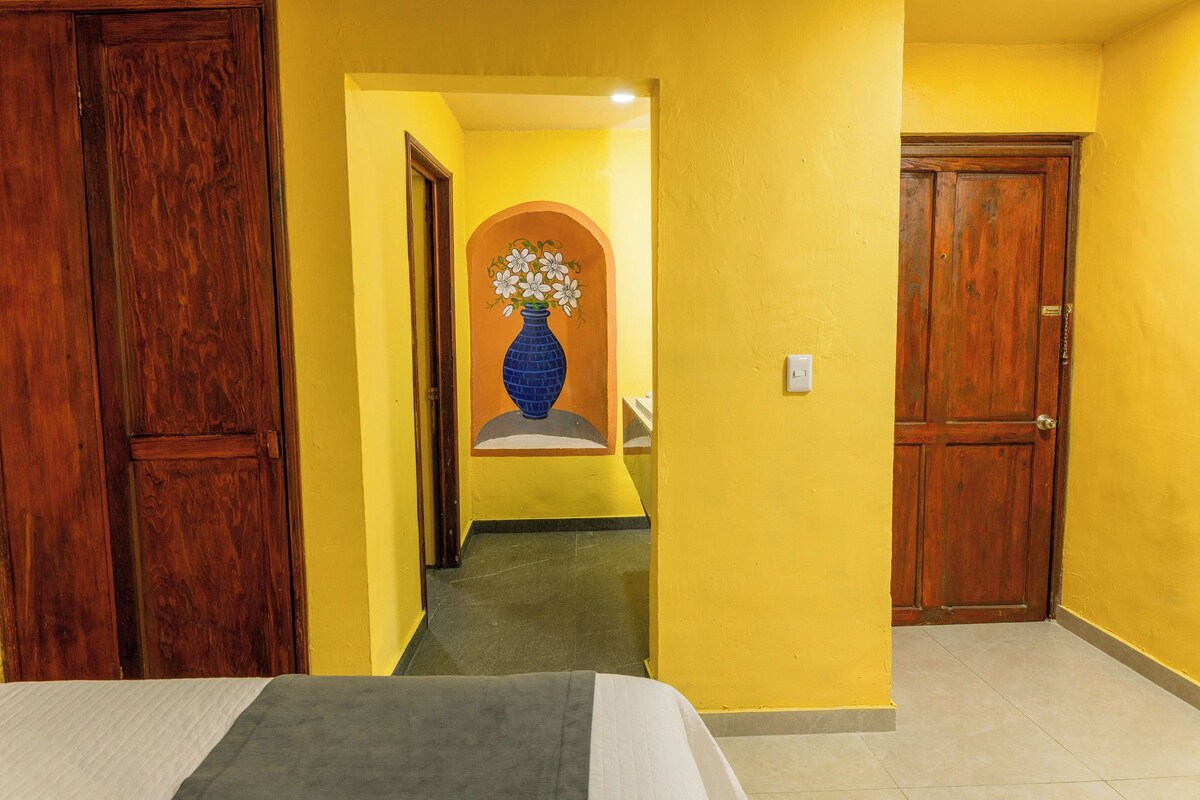 Un pasillo de hotel con puertas de madera, una pared amarilla y un jarrón azul con flores sobre un pedestal.