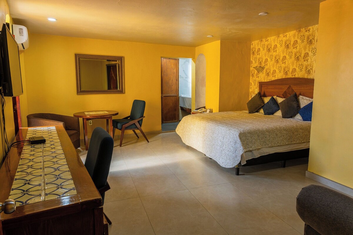 Une chambre d’hôtel comprenant un lit, un bureau, une chaise, un miroir et une porte donnant sur une autre pièce.