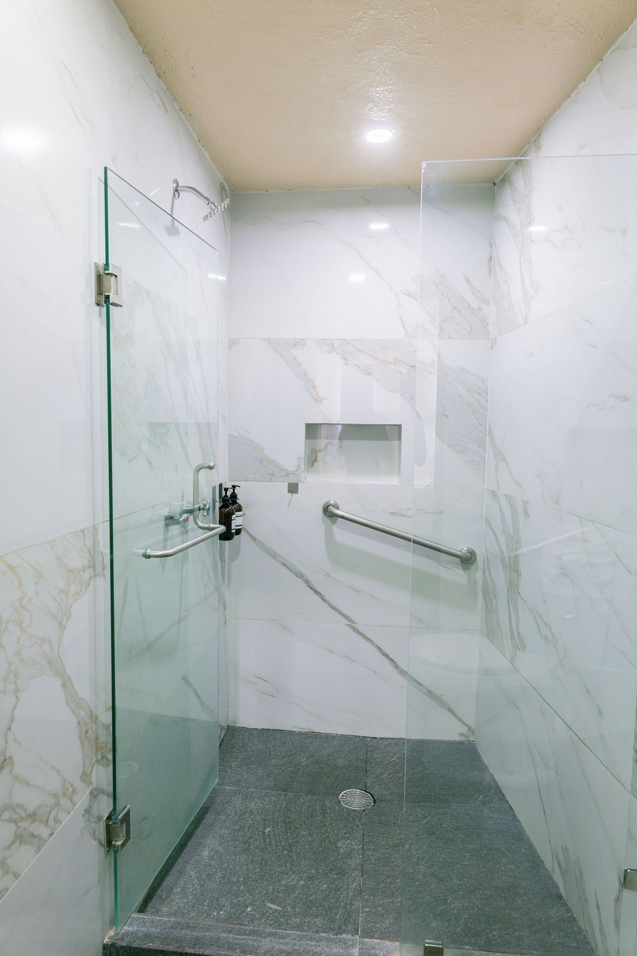 A glass-enclosed shower stall with a marble wall, a handrail, and a small shelf.