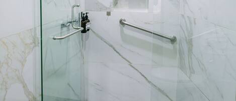 A glass-enclosed shower stall with a marble wall, a handrail, and a small shelf.
