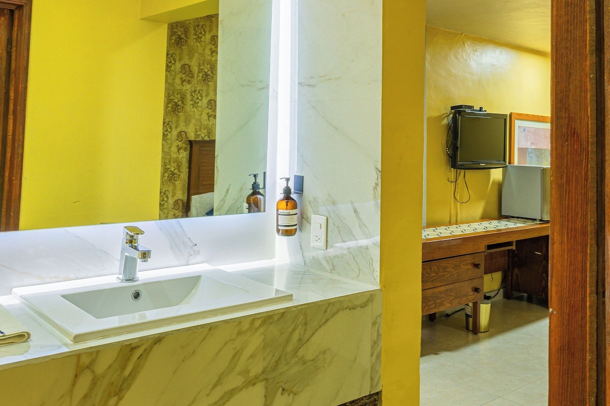 A bathroom with a marble sink, yellow walls, a mirror, a television, and a wooden cabinet.
