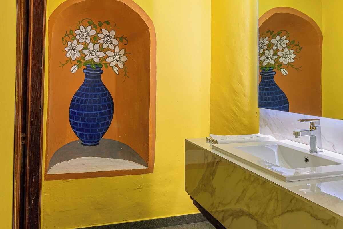 A bathroom with yellow walls, a marble sink, and a mirror reflecting a painted vase.
