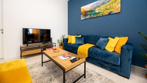 Een moderne woonkamer met een flatscreen televisie, een houten salontafel en een blauwe bank met gele kussens.