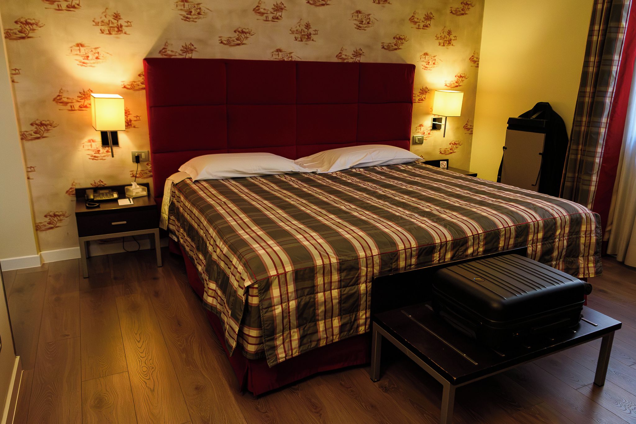 A hotel room with a bed, bedside tables, a lamp, a suitcase, and a mirror.