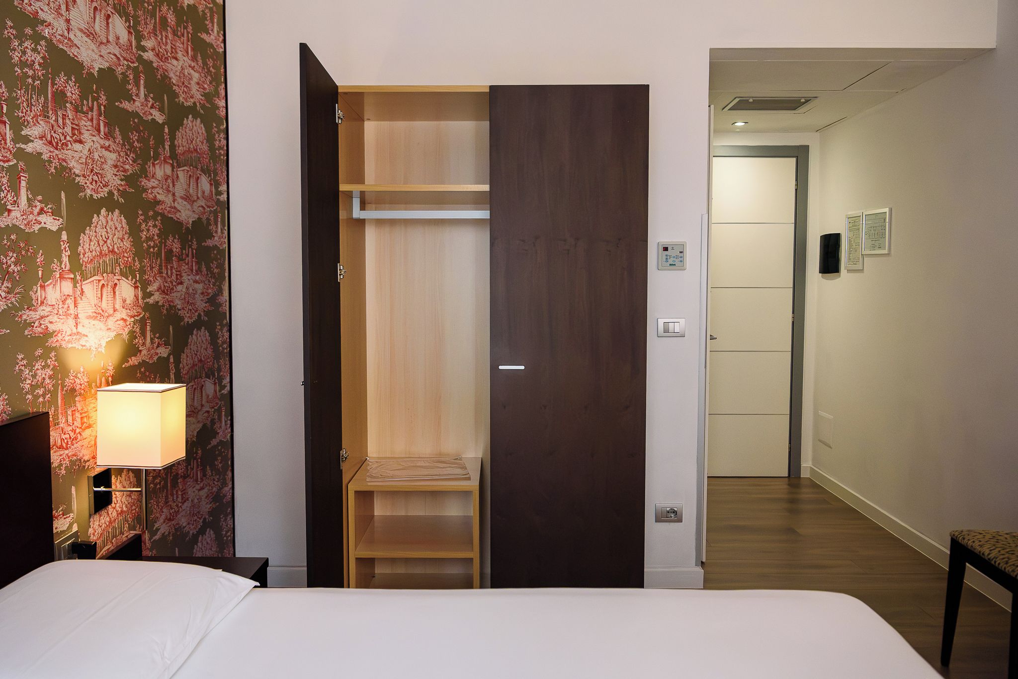 A hotel room with a bed, a nightstand, a lamp, a wardrobe, and a chair.