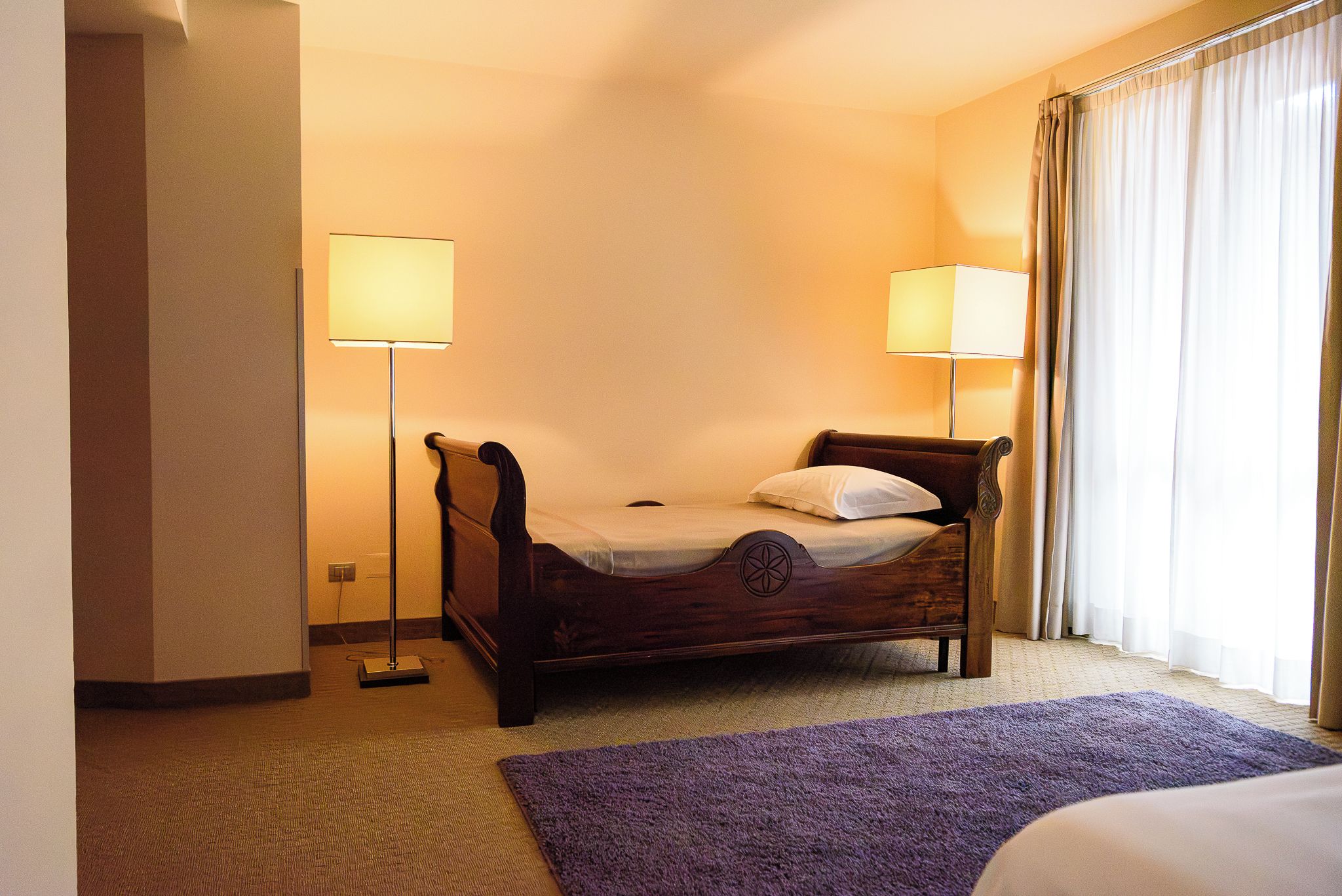 A hotel room with a single bed, two bedside lamps, a purple rug, and a window with curtains.