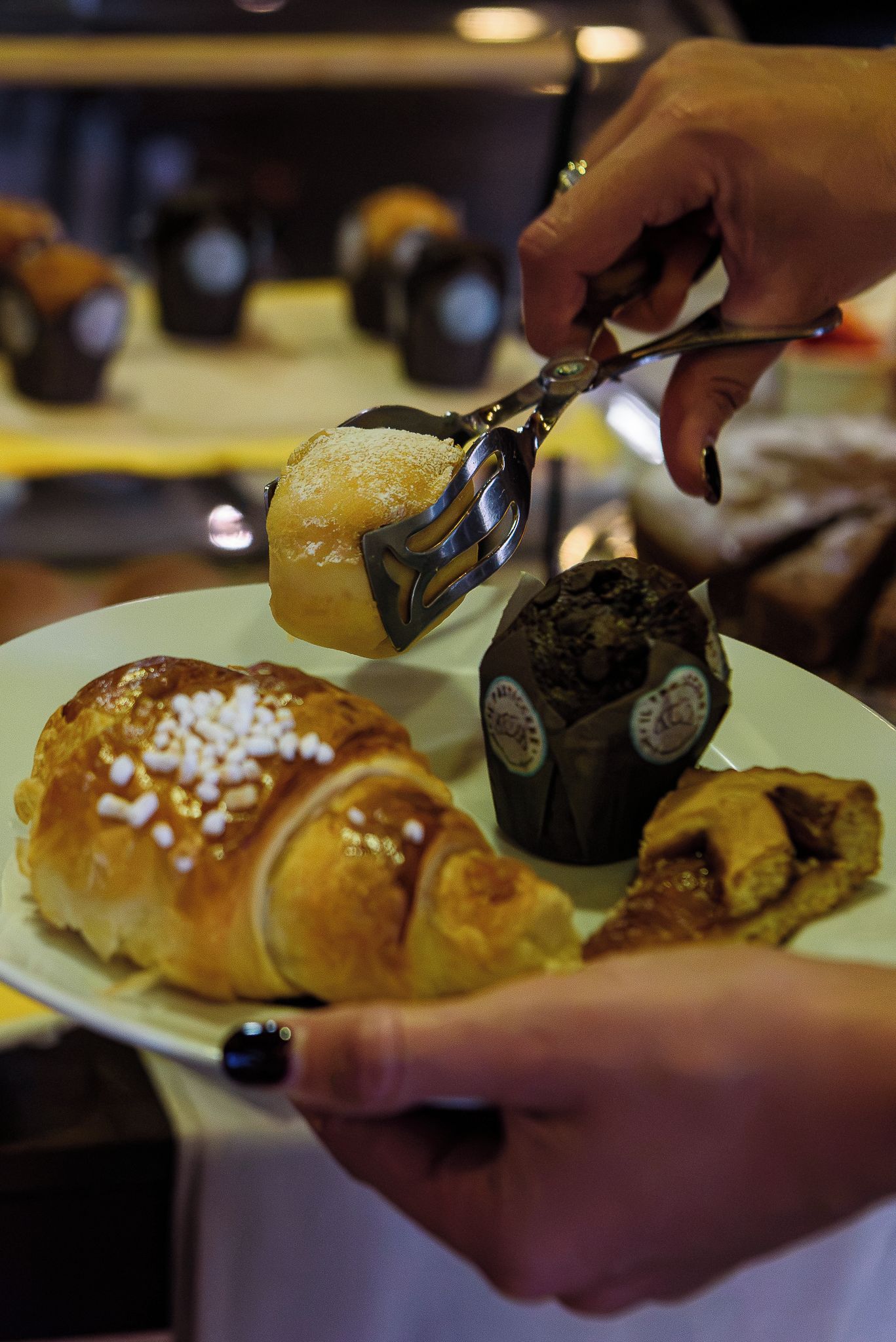 A person holding a pastry with a fork, a croissant, and a chocolate muffin on a plate.