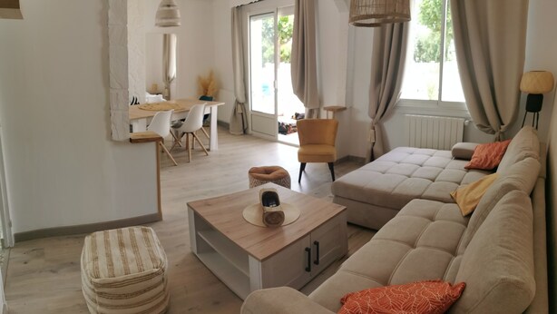 A modern living room with a beige sofa, a wooden coffee table, and a striped ottoman. There is a dining area with a table and chairs, and a window with curtains. - Maison à 5 Minutes de la Plage, la Chicane, Laiguillon sur Mer (L'Aiguillon-la-Presqu'île)