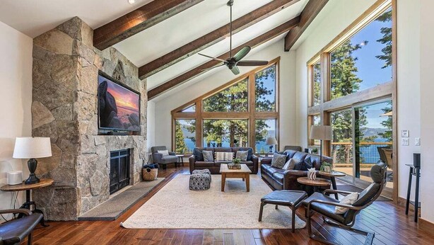 A spacious living room with a stone fireplace, large windows, and a high ceiling with exposed beams.