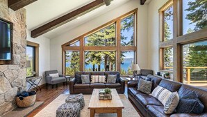A spacious living room with a stone fireplace, large windows offering a view of trees and a lake, and a cozy seating area with a brown leather sofa and patterned ottoman.