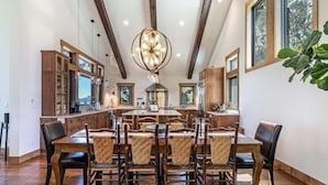 A spacious dining area with a large wooden table surrounded by chairs, a modern chandelier, and wooden cabinets.