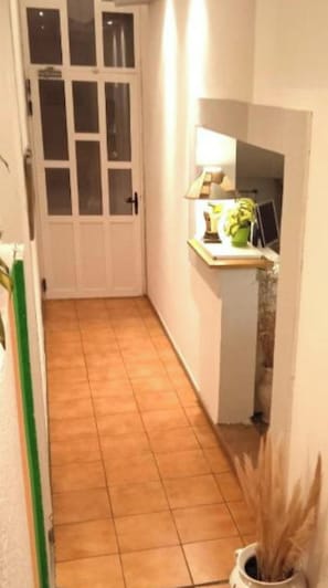 Un couloir étroit au sol carrelé, une porte blanche avec des panneaux de verre, et un petit comptoir sur lequel se trouvent une lampe et une plante.