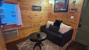 Tunnelmallinen, mökin tyylinen huone, jossa on puuseinät, musta nahkasohva, pieni pyöreä pöytä ja seinälle kiinnitetty televisio.