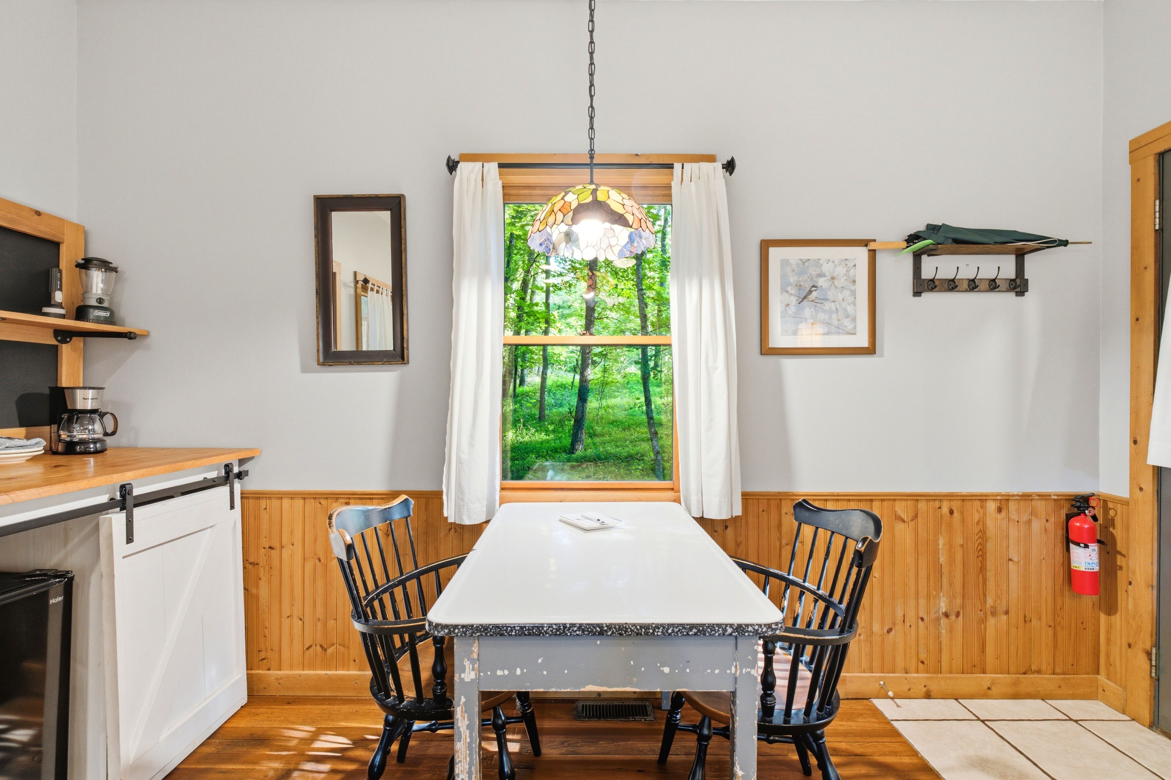 Et spiseområde med et bord og stole, et vindue med udsigt over grøn vegetation, en loftlampe og en brandslukker.